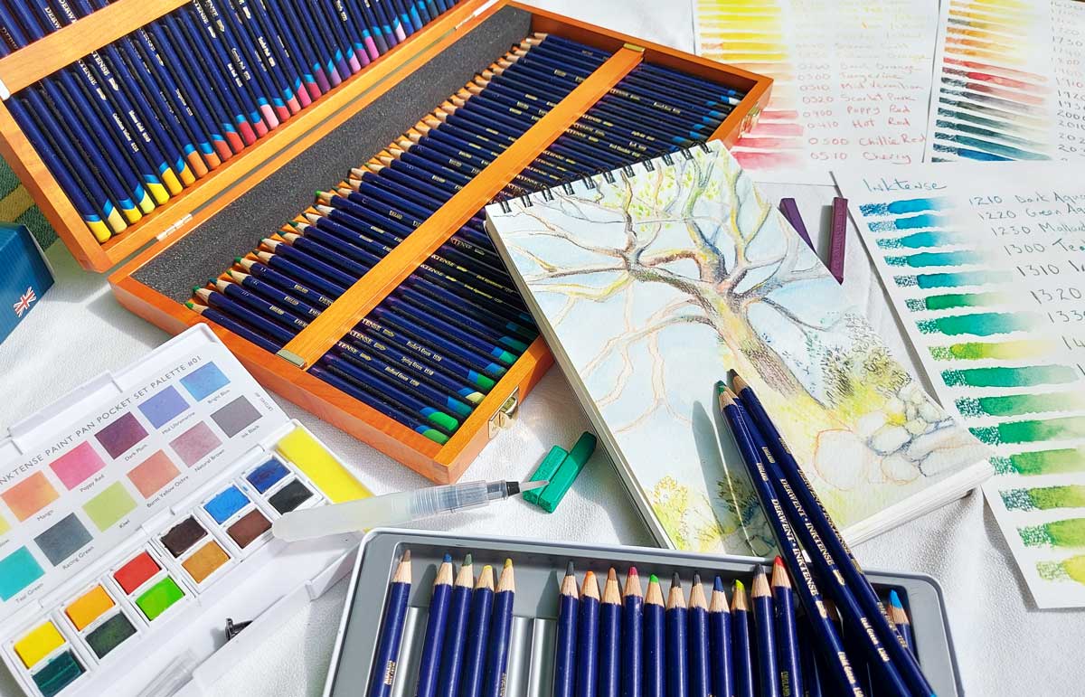 A watercolour pencil image of a tree in a white A5 sketchbook resting on a wooden box full of Inktense coloured pencils.