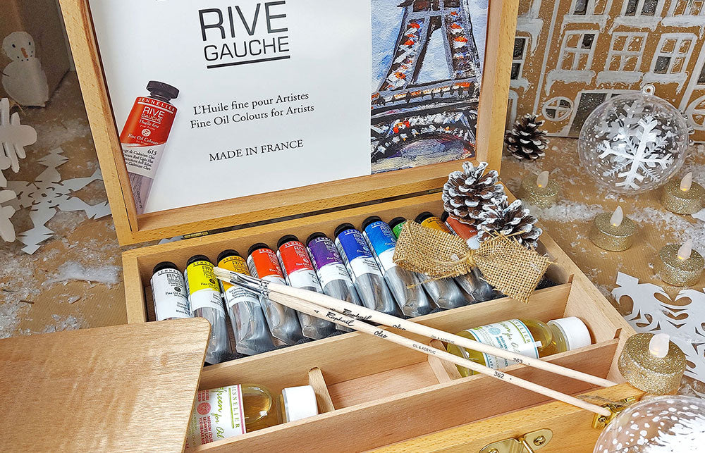 Open wooden box full of Sennelier Rive Gauche Oil paints with two Raphael paint brushes laying over the tubes of paint, Painted pine cones and scattered artificial snow surround the box..