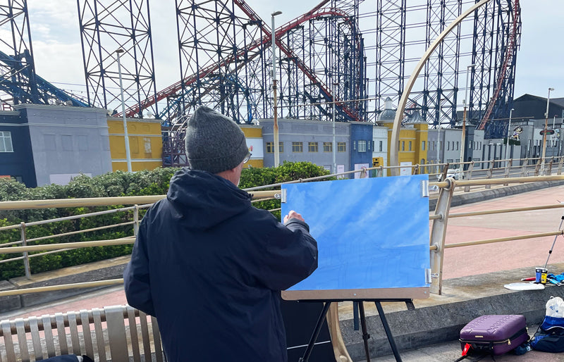 Artist Alan Fernihough standing in front of his easel with his back to the camera and looking over his easel at Blackpool Pleasure Beach roller-coaster.