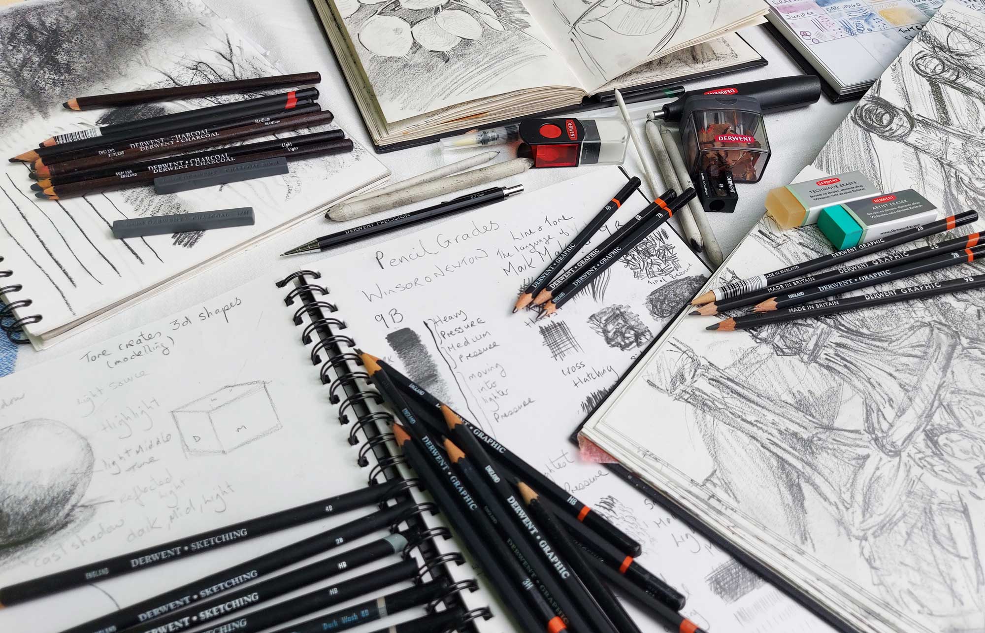 Open sketchbooks with drawings studies and drawings of bottles by Ellie Jakeman. Derwent Graphic pencils scattered over the pages.