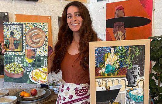 Artist Donna Flowers-Dorning stood in front of a wall covered in her vibrant paintings, she is holding two still life paintings one in each hand and is wearing a patterned skirt.