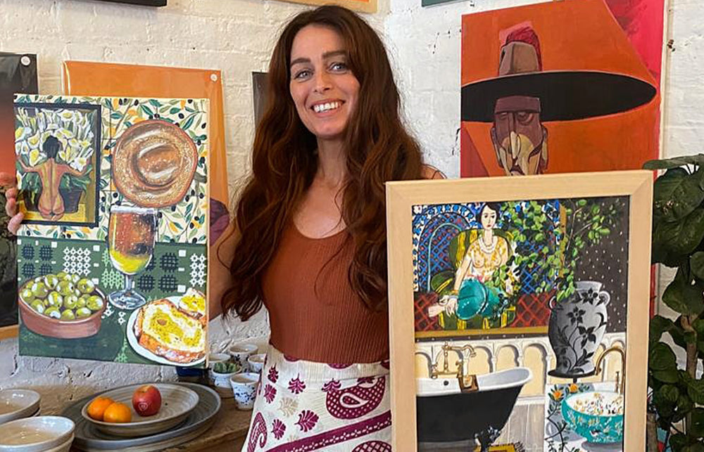 Artist Donna Flowers-Dorning stood in front of a wall covered in her vibrant paintings, she is holding two still life paintings one in each hand and is wearing a patterned skirt.
