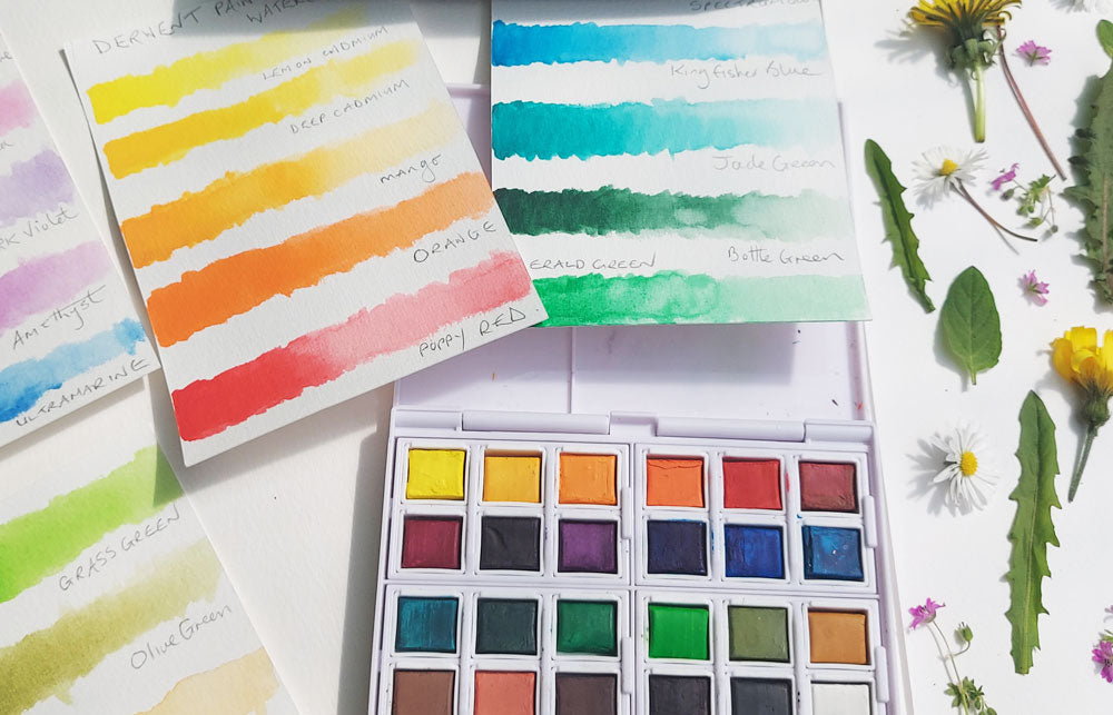 Inktense half pan watercolour set open on a white piece of paper covered in dandelion and daisy flower heads and leaves with sample colour charts.