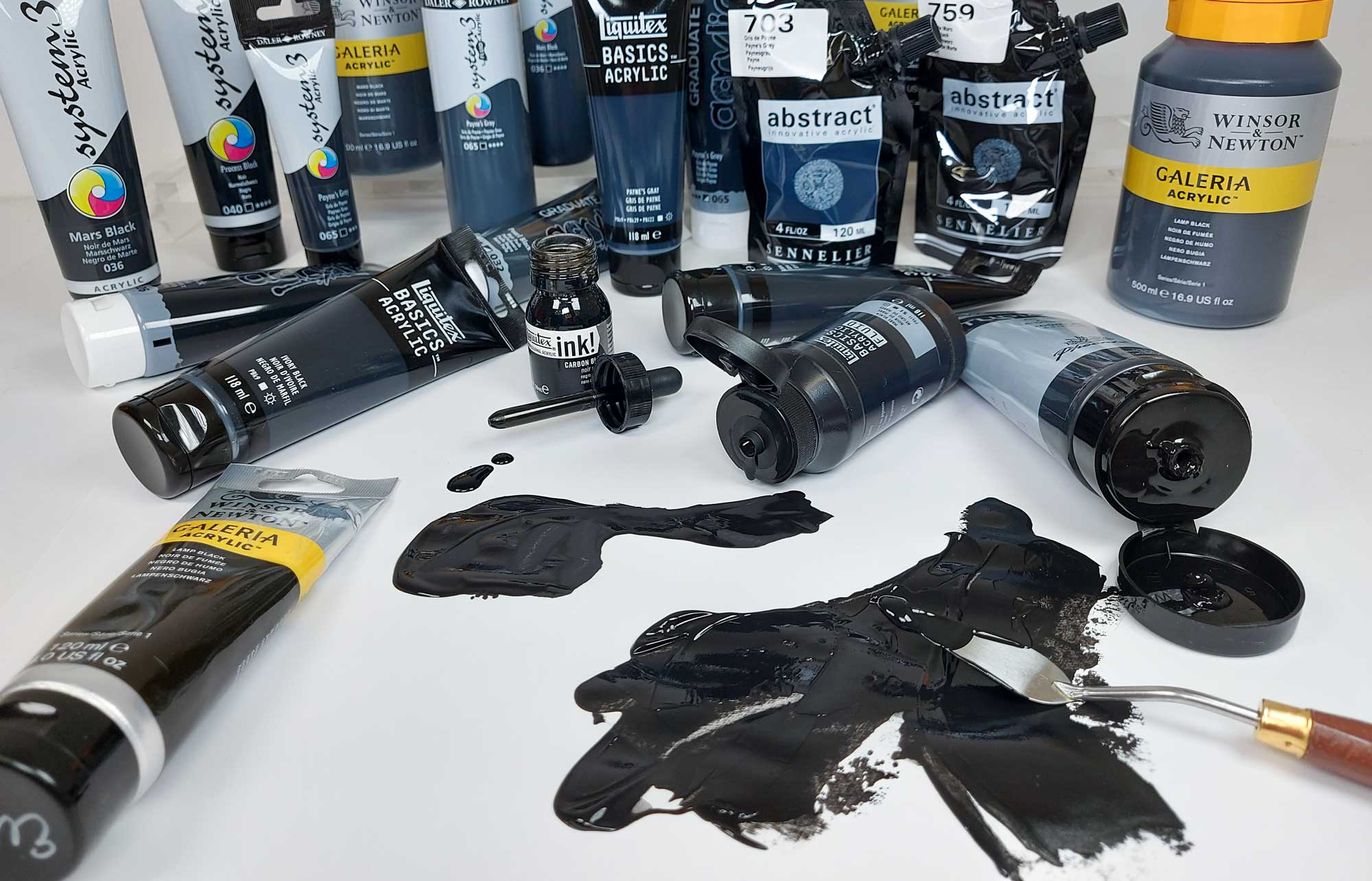 Lots of black artists products scattered on a white desk. Bottles of black ink, tubes of black paint, 