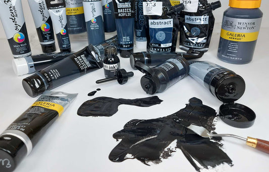 Lots of black artists products scattered on a white desk. Bottles of black ink, tubes of black paint, 