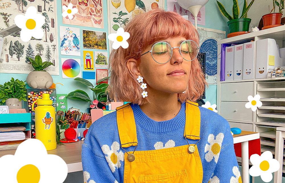 Artist Ivna Lins sat in front of a wall displaying all of her colourful illustrations in her studio. She wears a daisy earrings and a daisy patterned Jumper. 