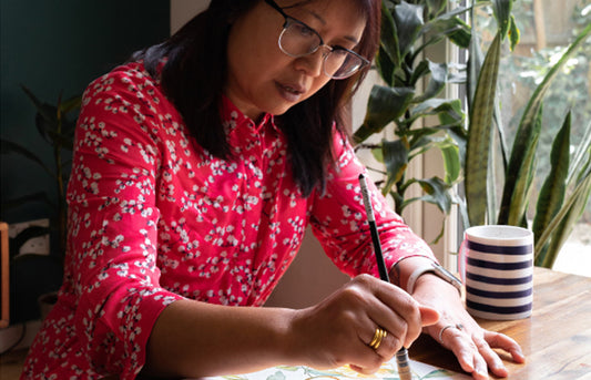Artist Ohn Mar Win, sat her desk with a paintbrush in her right hand. she is wearing a bright pink with tiny white flowers on it.