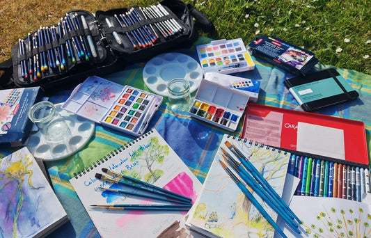 image of a sketchpad and brushes and water colour sets on a blanket outside. 