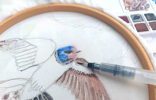 Embroidered  swallow drawing in an embroidery hoop with a waterbrush wetting an area coloured in with Inktense pencils.