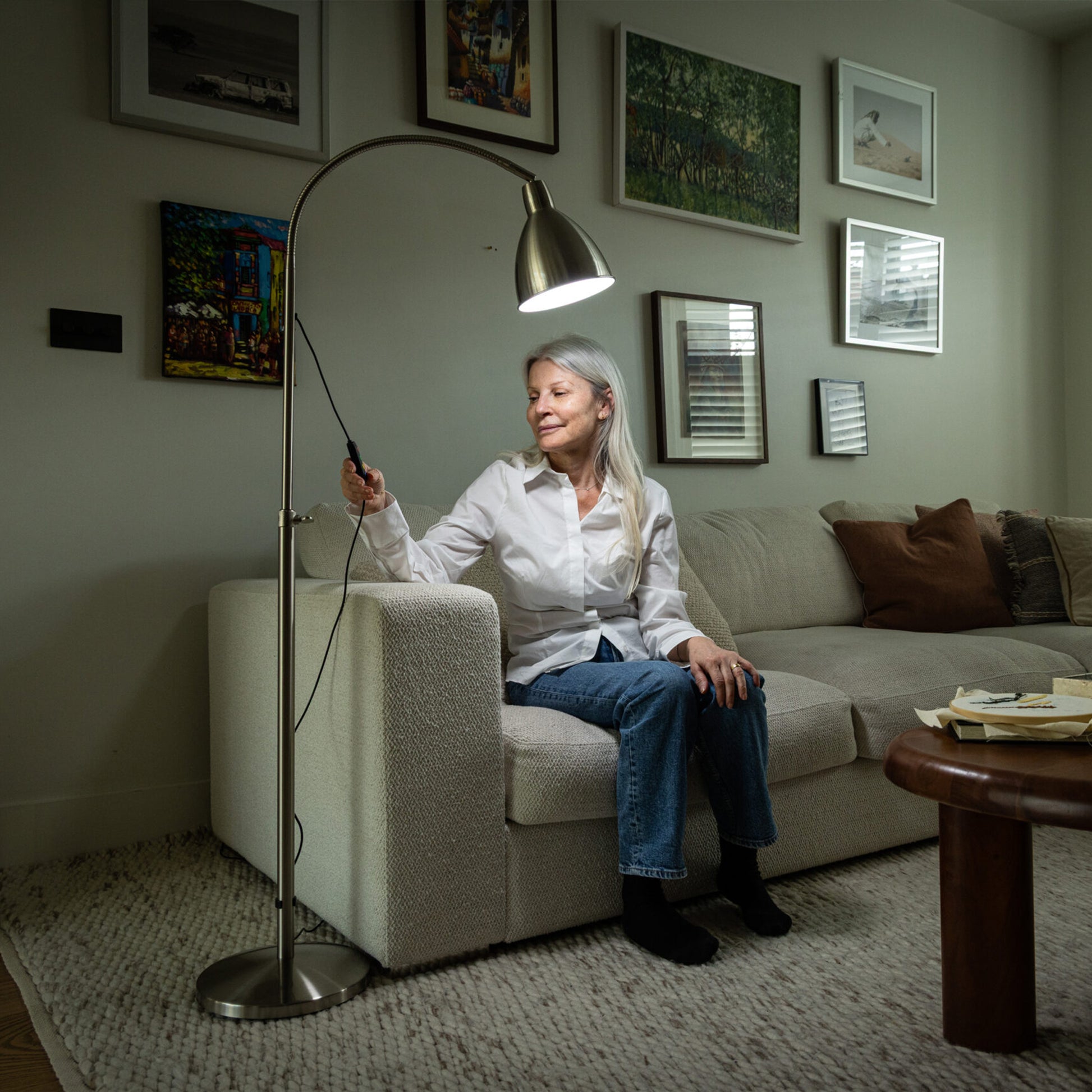 Woman sitting on a couch in a living room adjusting a floor lamp.