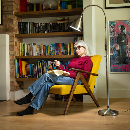 Woman reading a book in a yellow chair in a room with bookshelves and a lamp.